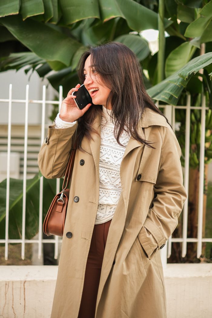 Stylish woman in trench coat happily talking on smartphone outdoors.
